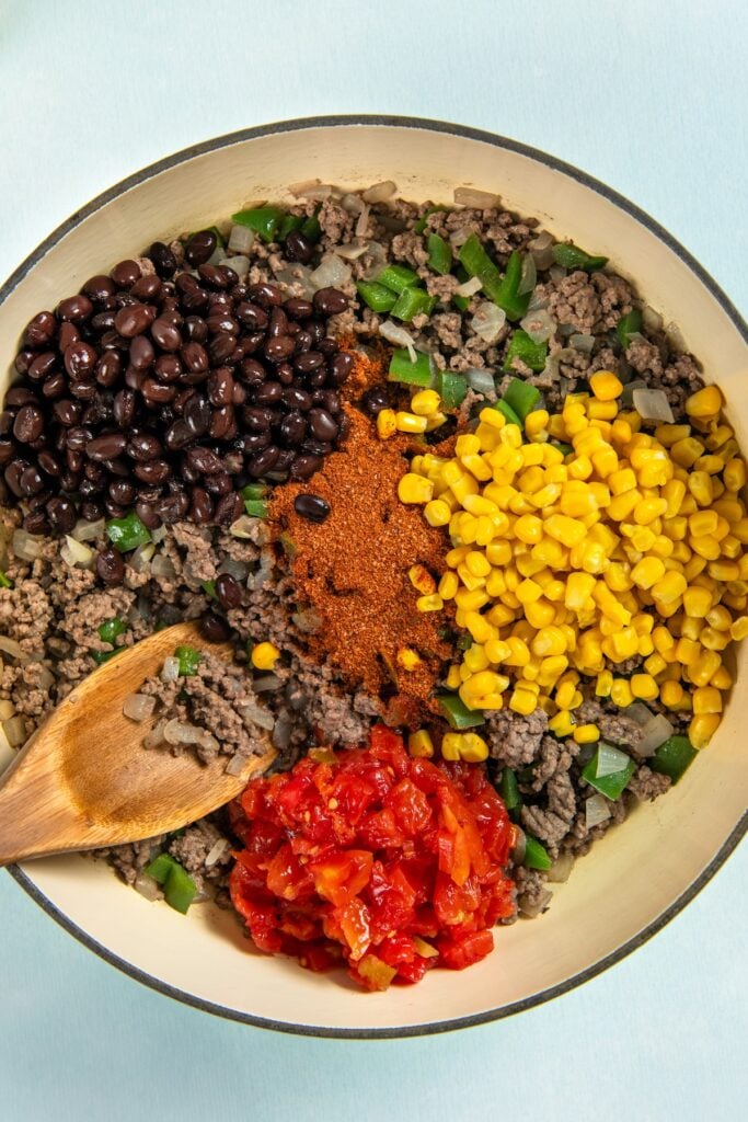 This hearty and cheesy Mexican Beef and Rice Skillet is an easy one‑pan dinner that's ready in just about 30 minutes. Ultimate comfort food! A skillet filled with cooked ground beef, black beans, corn, diced tomatoes, green bell peppers, and chili powder, with a wooden spoon resting inside. Ingredients are arranged in sections and not yet mixed.