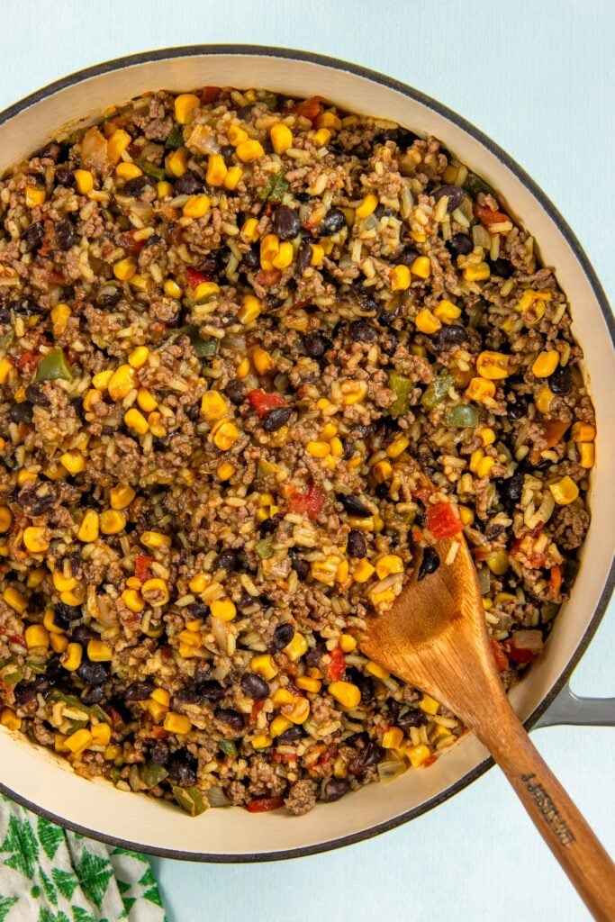 This hearty and cheesy Mexican Beef and Rice Skillet is an easy one‑pan dinner that's ready in just about 30 minutes. Ultimate comfort food! A close-up of a large pot filled with a colorful mixture of rice, black beans, corn, diced tomatoes, and bell peppers, with a wooden spoon resting inside.