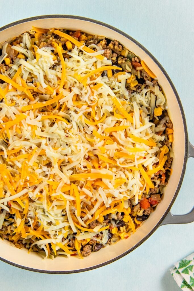 This hearty and cheesy Mexican Beef and Rice Skillet is an easy one‑pan dinner that's ready in just about 30 minutes. Ultimate comfort food! A large skillet filled with a mixture of ground meat, beans, corn, and vegetables, all topped with a generous layer of shredded yellow and white cheese, ready to be baked.