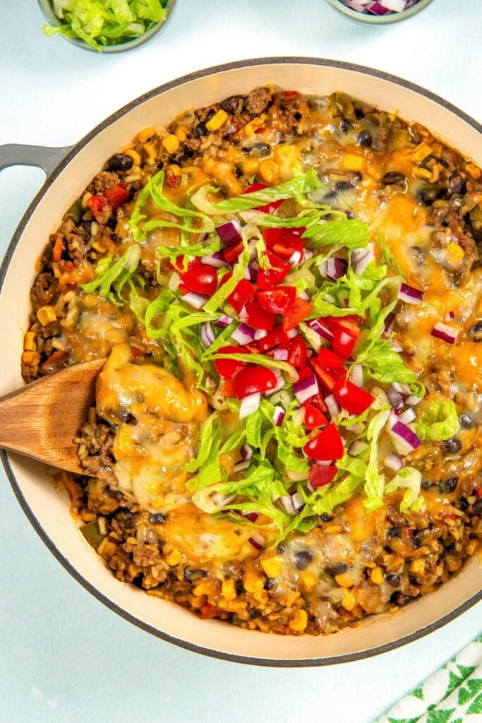 This hearty and cheesy Mexican Beef and Rice Skillet is an easy one‑pan dinner that's ready in just about 30 minutes. Ultimate comfort food! A large skillet filled with cheesy Mexican beef and rice skillet, topped with shredded lettuce, diced tomatoes, and chopped red onion. A wooden spoon rests in the skillet, ready to serve.