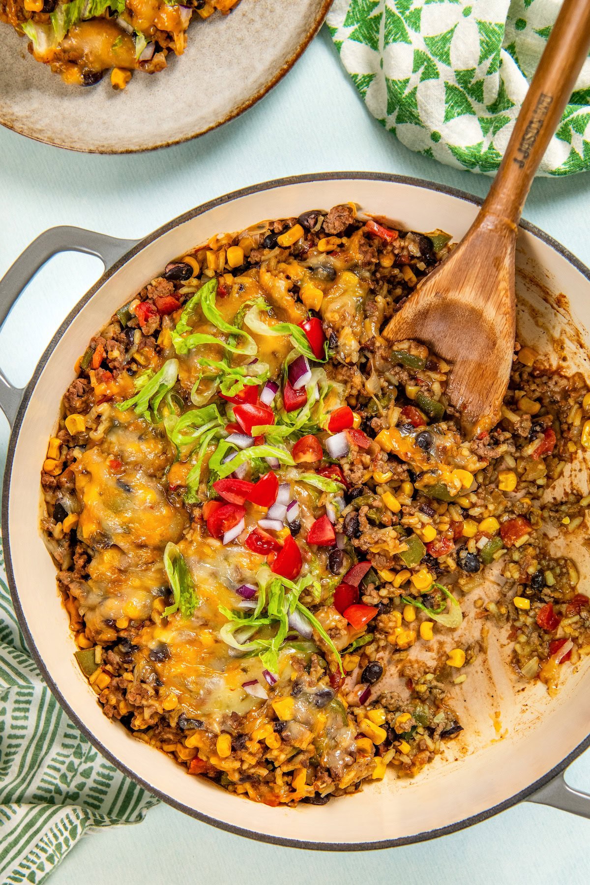 This hearty and cheesy Mexican Beef and Rice Skillet is an easy one‑pan dinner that's ready in just about 30 minutes. Ultimate comfort food! A pot filled with a colorful Mexican beef and rice skillet topped with melted cheese, diced tomatoes, red onion, and sliced lettuce. A few servings have been scooped out and a wooden spoon rests in the skillet.