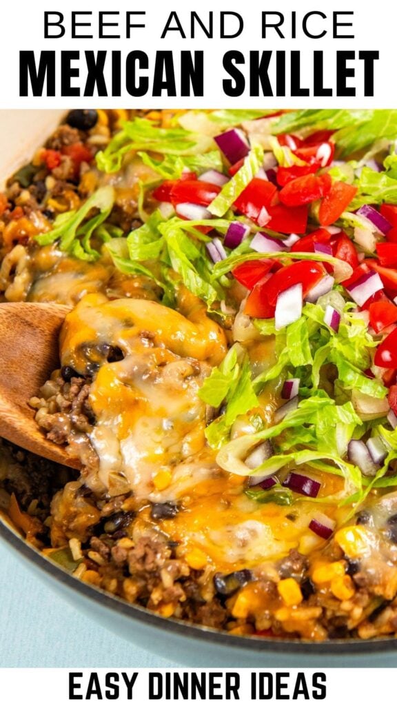 A close-up of a beef and rice Mexican beef and rice skillet topped with melted cheese, chopped lettuce, tomatoes, and red onions. A wooden spoon scoops a serving.