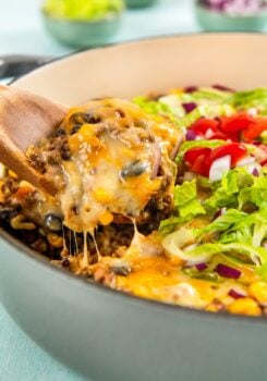 A wooden spoon lifts a serving of gooey, cheesy Mexican beef and rice skillet from a pot. The dish is topped with chopped lettuce, tomatoes, and red onions.