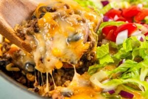 A wooden spoon lifts a serving of gooey, cheesy Mexican beef and rice skillet from a pot. The dish is topped with chopped lettuce, tomatoes, and red onions.