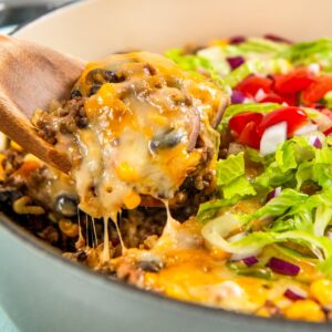 A wooden spoon lifts a serving of gooey, cheesy Mexican beef and rice skillet from a pot. The dish is topped with chopped lettuce, tomatoes, and red onions.