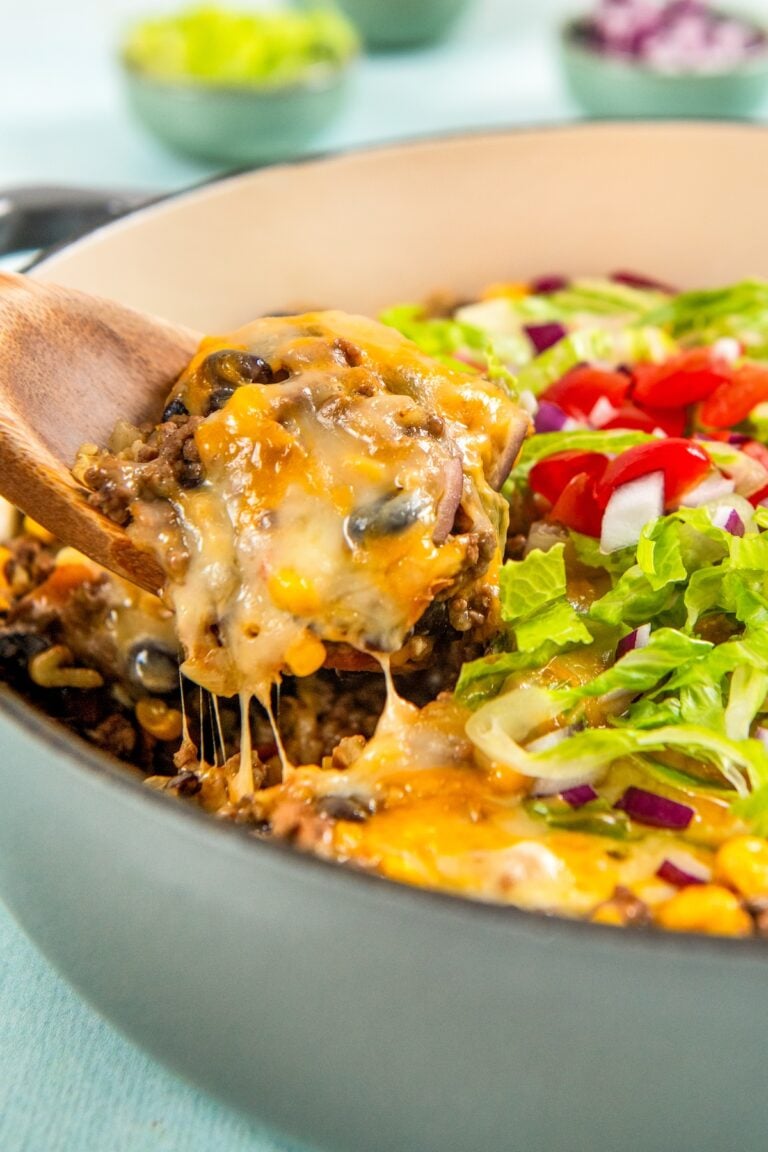 A wooden spoon lifts a serving of gooey, cheesy Mexican beef and rice skillet from a pot. The dish is topped with chopped lettuce, tomatoes, and red onions.