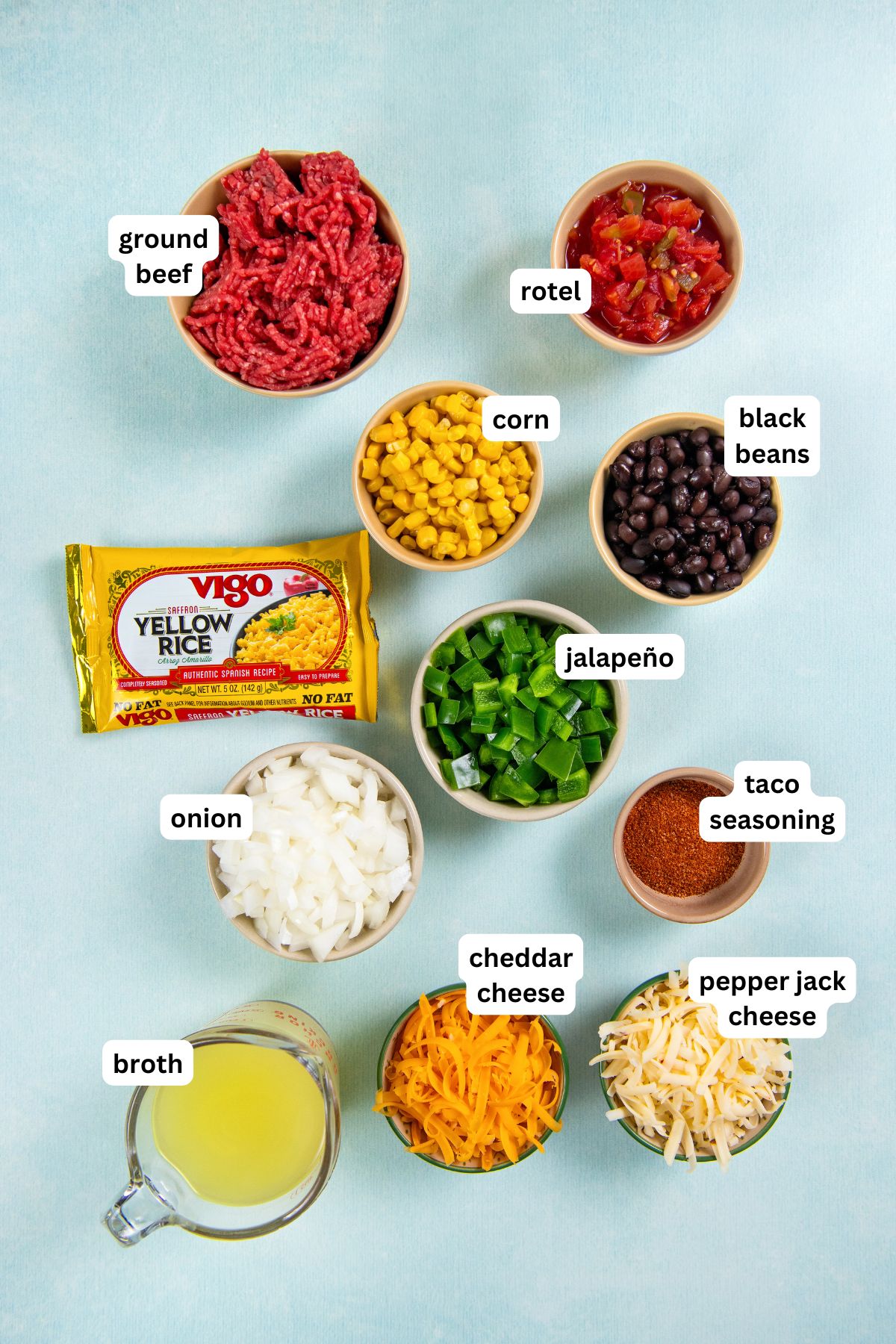 This hearty and cheesy Mexican Beef and Rice Skillet is an easy one‑pan dinner that's ready in just about 30 minutes. Ultimate comfort food! Ingredients for Mexican Skillet recipe arranged on a light blue surface, including labeled bowls of ground beef, Rotel tomatoes, corn, black beans, jalapeño, diced onion, taco seasoning, cheddar cheese, pepper jack cheese, yellow rice, and a measuring cup of broth.