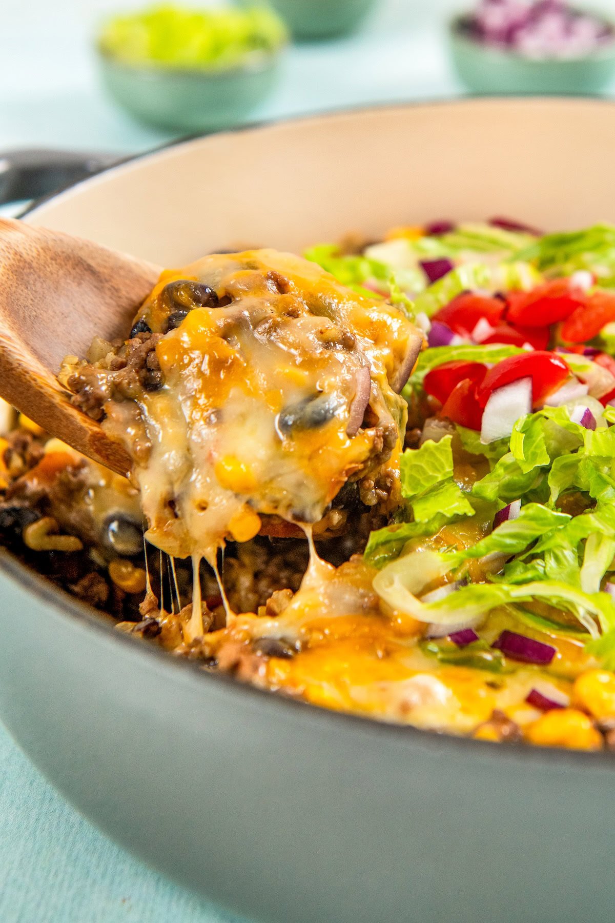 This hearty and cheesy Mexican Beef and Rice Skillet is an easy one‑pan dinner that's ready in just about 30 minutes. Ultimate comfort food! A wooden spoon lifts a serving of gooey, cheesy Mexican beef and rice skillet from a pot. The dish is topped with chopped lettuce, tomatoes, and red onions.