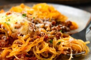 A plate of spaghetti squash topped with meat sauce, melted cheese, and herbs, with a fork twirling some of the squash.