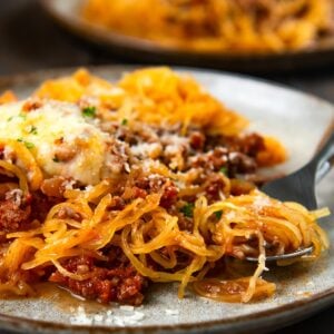 A plate of spaghetti squash topped with meat sauce, melted cheese, and herbs, with a fork twirling some of the squash.