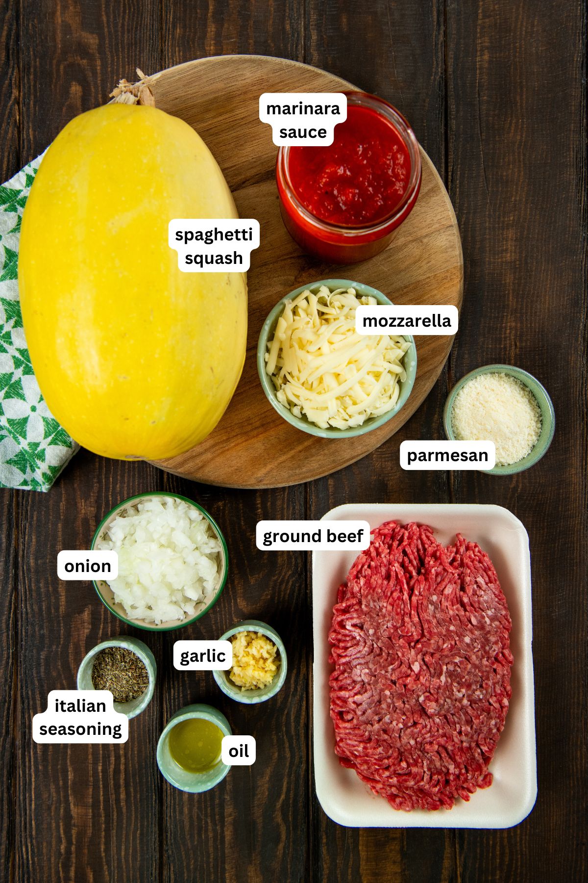 Stuffed spaghetti squash filled with a saucy ground meat mixture and gooey melted cheese. A hearty, low-carb comfort food with a veggie twist. Ingredients for a stuffed spaghetti squash recipe are arranged on a dark wooden surface, including a whole spaghetti squash, marinara sauce, shredded mozzarella, grated parmesan, ground beef, chopped onion, minced garlic, Italian seasoning, and oil.