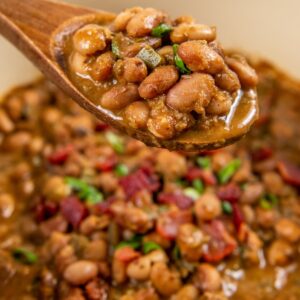 A close-up of a wooden spoon lifting a portion of cooked pinto beans in a thick, savory sauce from a pot topped with crispy bacon and sliced green onions.
