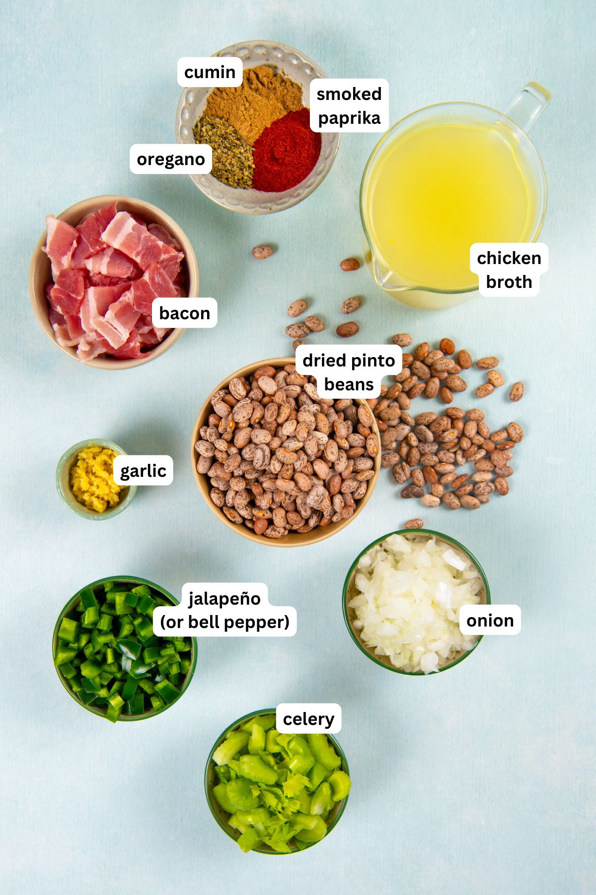Learn how to cook pinto beans to tender and creamy perfection in a smoky, flavorful broth with bacon, jalapeño, onion, and more. Overhead view of ingredients for dried pinto beans recipe, including labeled bowls of cumin, smoked paprika, oregano, bacon, chicken broth, dried pinto beans, garlic, jalapeño, onion, and celery.
