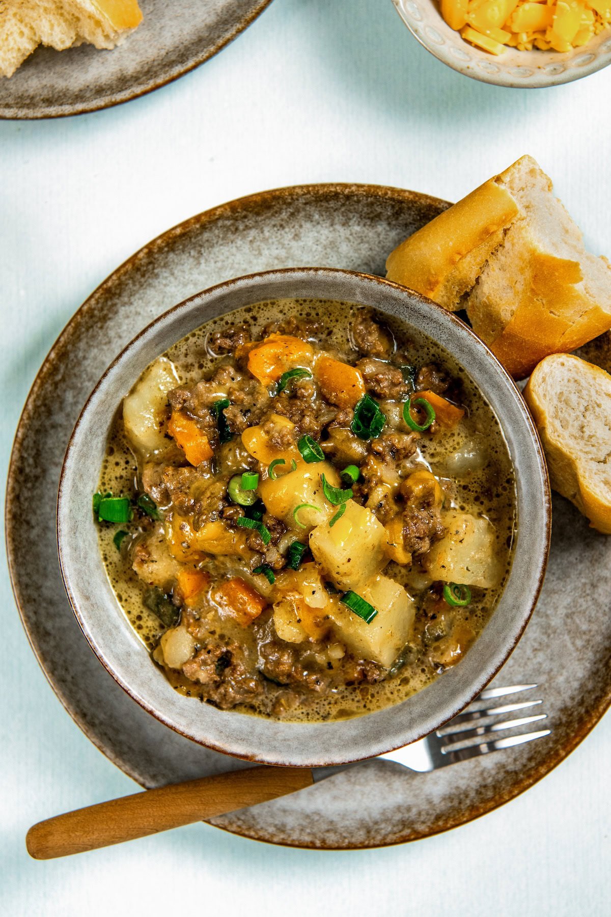 Ground beef, potatoes, and plenty of cheese combine to make this Hamburger Hash recipe, an easy one-pot dinner that's ready in 30 minutes! A bowl of hearty hamburger hash with ground beef, potatoes, carrots, and green onions, served with slices of baguette on a gray plate.