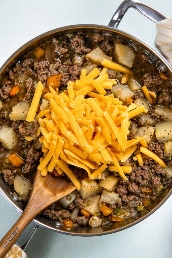 Ground beef, potatoes, and plenty of cheese combine to make this Hamburger Hash recipe, an easy one-pot dinner that's ready in 30 minutes! A skillet filled with cooked ground beef, diced potatoes, carrots, and onions, topped with a generous pile of shredded cheddar cheese. A wooden spoon rests in the skillet, ready to stir the ingredients.