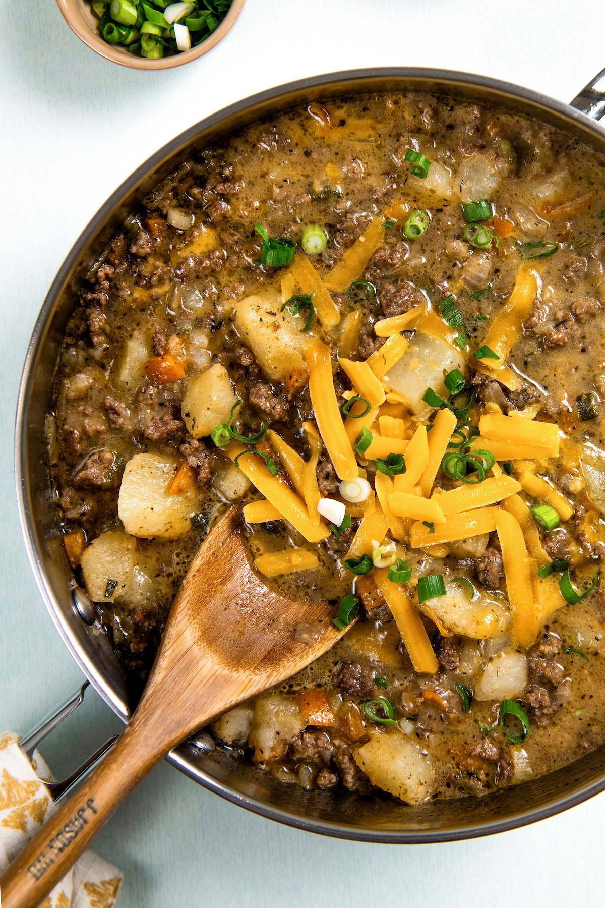 Ground beef, potatoes, and plenty of cheese combine to make this Hamburger Hash recipe, an easy one-pot dinner that's ready in 30 minutes! A skillet filled with a cheesy hamburger hash with potatoes, topped with shredded cheddar cheese and chopped green onions. A wooden spoon rests inside the pan, and a small bowl of sliced green onions is nearby.