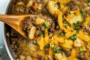 A skillet filled with cheesy hamburger hash with ground beef and potatoes, topped with shredded cheddar cheese and chopped green onions. A wooden spoon is scooping up a portion for serving.