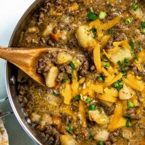 A skillet filled with cheesy hamburger hash with ground beef and potatoes, topped with shredded cheddar cheese and chopped green onions. A wooden spoon is scooping up a portion for serving.