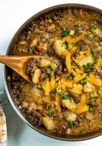 A skillet filled with cheesy hamburger hash with ground beef and potatoes, topped with shredded cheddar cheese and chopped green onions. A wooden spoon is scooping up a portion for serving.