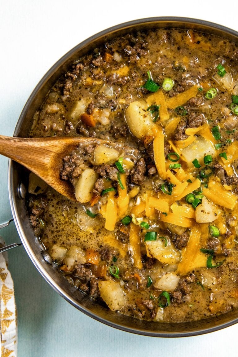 A skillet filled with cheesy hamburger hash with ground beef and potatoes, topped with shredded cheddar cheese and chopped green onions. A wooden spoon is scooping up a portion for serving.