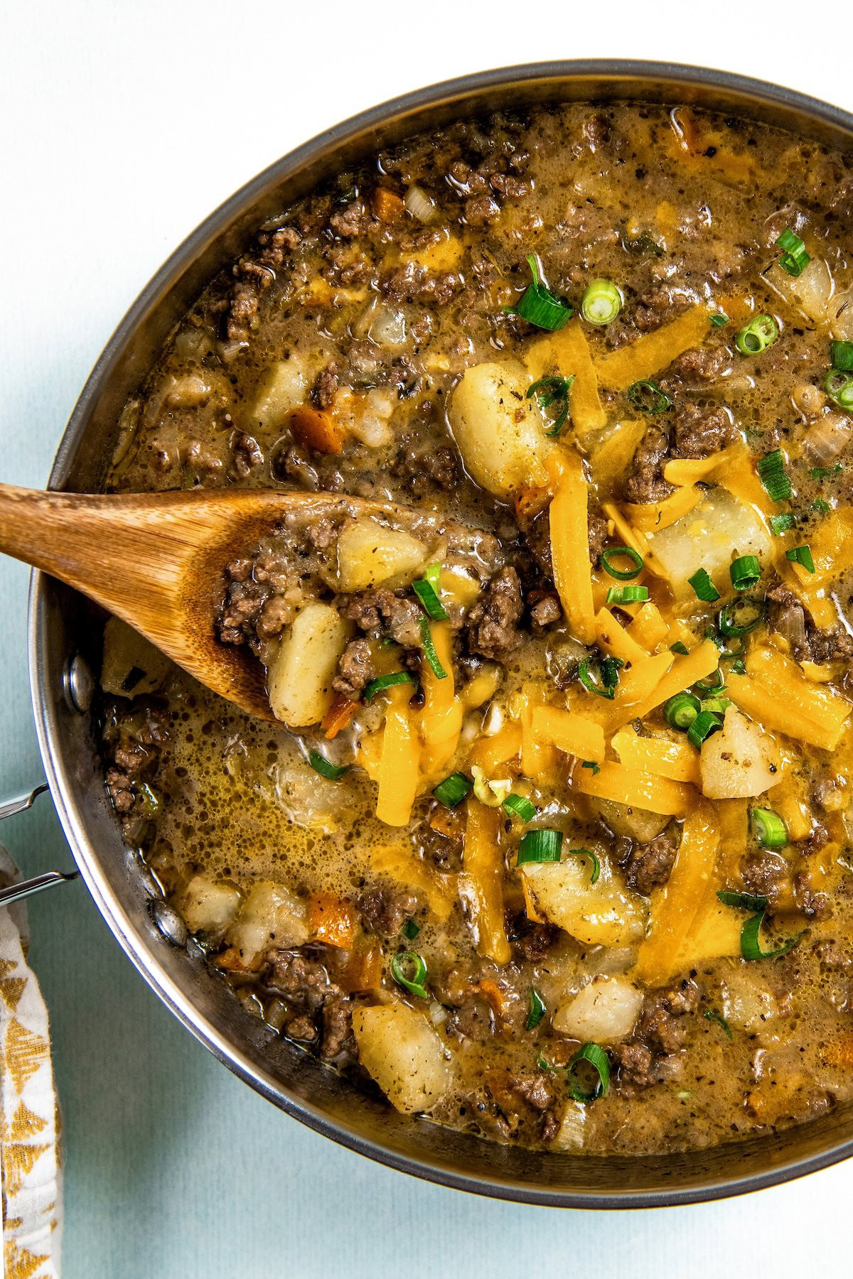 A skillet filled with cheesy hamburger hash with ground beef and potatoes, topped with shredded cheddar cheese and chopped green onions. A wooden spoon is scooping up a portion for serving.