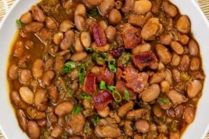 A bowl of seasoned pinto beans garnished with chopped green onions and bacon pieces, served on a yellow and white striped cloth with a small dish of cornbread in the background.