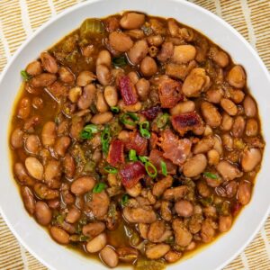 A bowl of seasoned pinto beans garnished with chopped green onions and bacon pieces, served on a yellow and white striped cloth with a small dish of cornbread in the background.