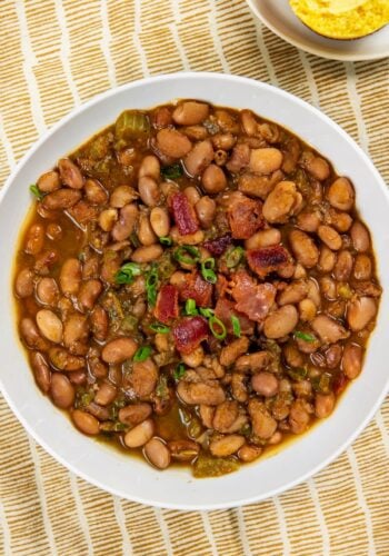 A bowl of seasoned pinto beans garnished with chopped green onions and bacon pieces, served on a yellow and white striped cloth with a small dish of cornbread in the background.