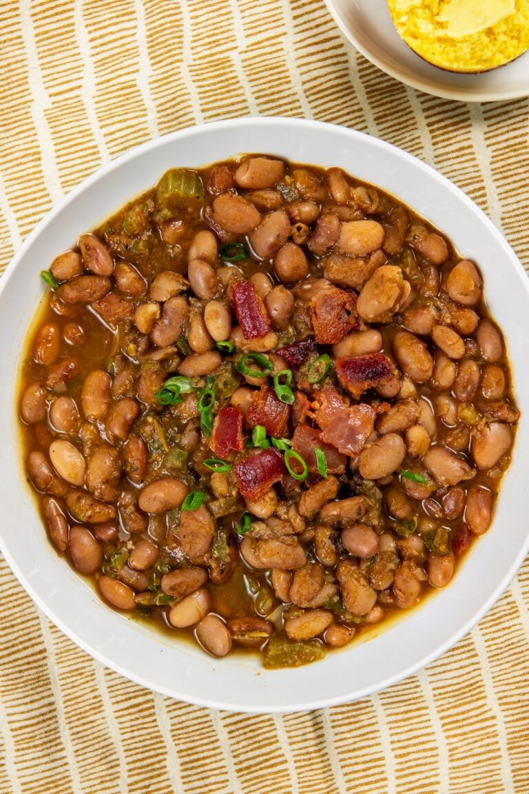 A bowl of seasoned pinto beans garnished with chopped green onions and bacon pieces, served on a yellow and white striped cloth with a small dish of cornbread in the background.