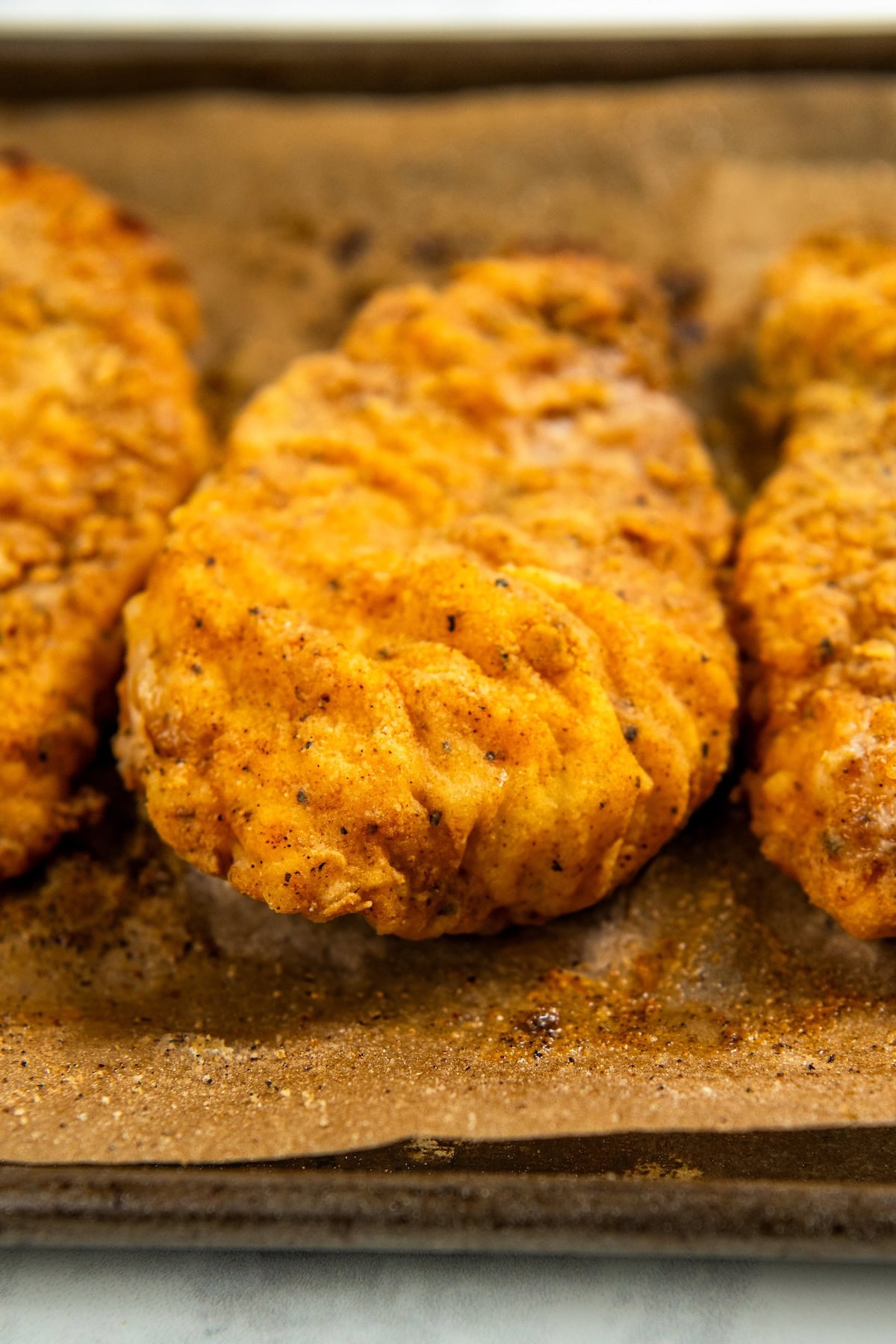 This Oven Fried Chicken recipe is always crispy, golden, perfectly juicy, and packed with flavor — without standing over a pot of hot oil. Close-up of three pieces of golden-brown, crispy oven baked fried chicken on a parchment-lined baking sheet, showing a crunchy, textured coating and seasoning.