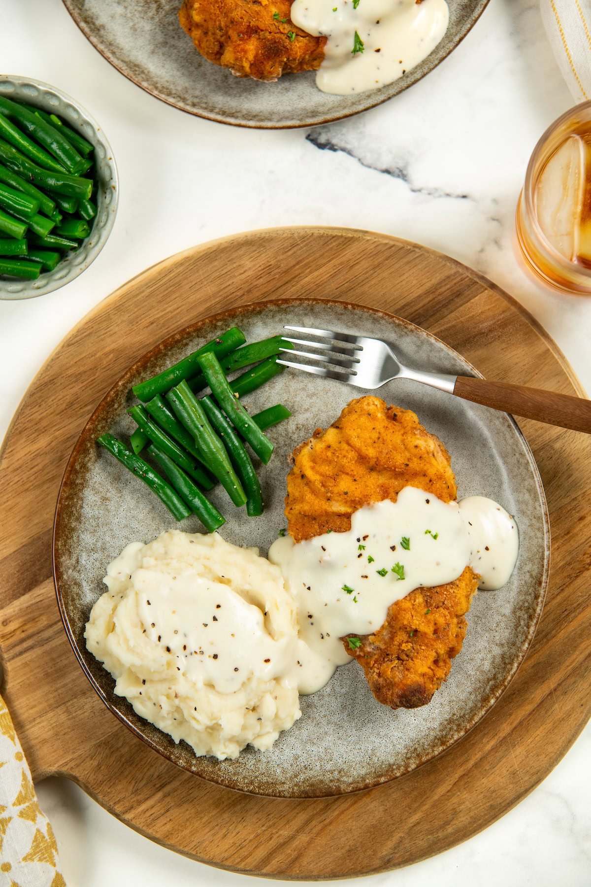 This Oven Fried Chicken recipe is always crispy, golden, perfectly juicy, and packed with flavor — without standing over a pot of hot oil. A plate with crispy oven baked fried chicken topped with white gravy with mashed potatoes and green beans on the side.