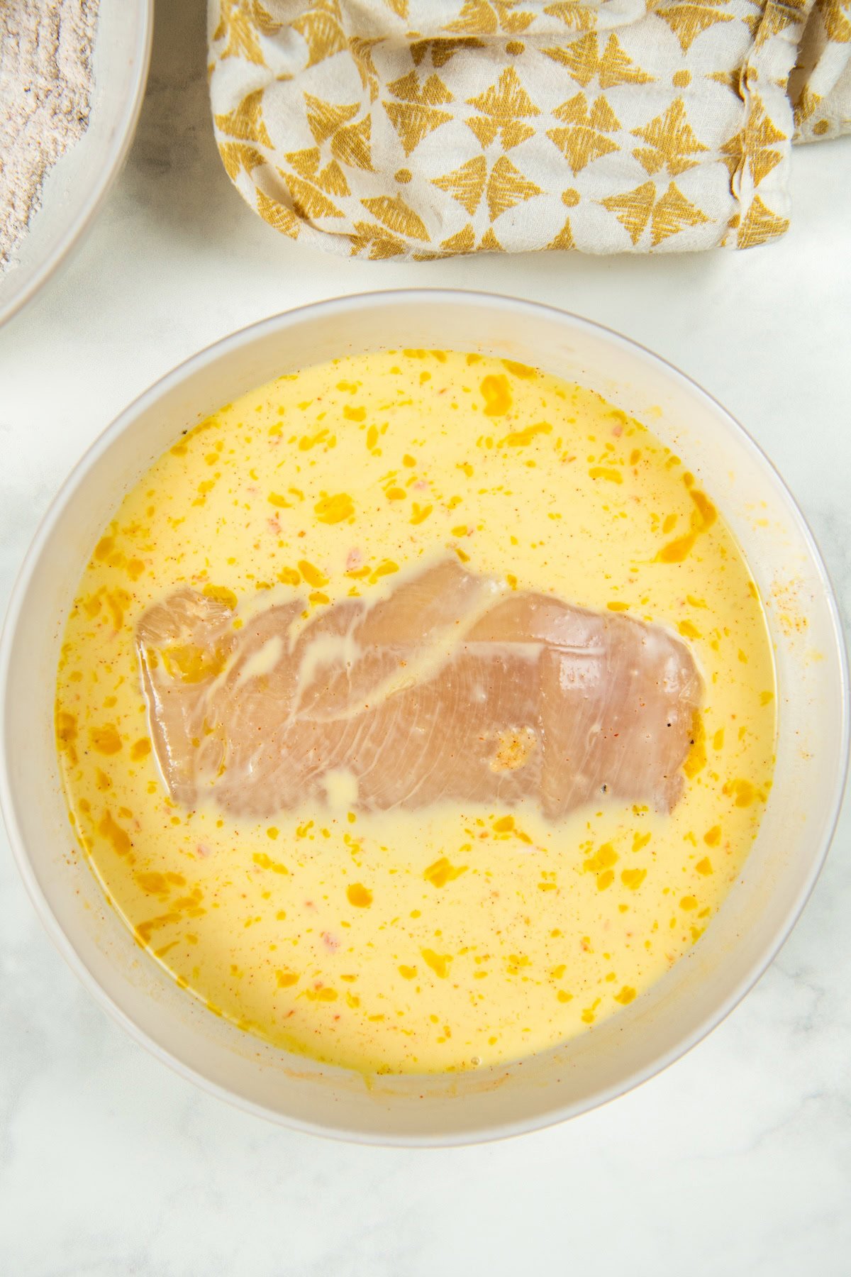 This Oven Fried Chicken recipe is always crispy, golden, perfectly juicy, and packed with flavor — without standing over a pot of hot oil. A raw chicken breast sits in a bowl of an egg and milk mixture.