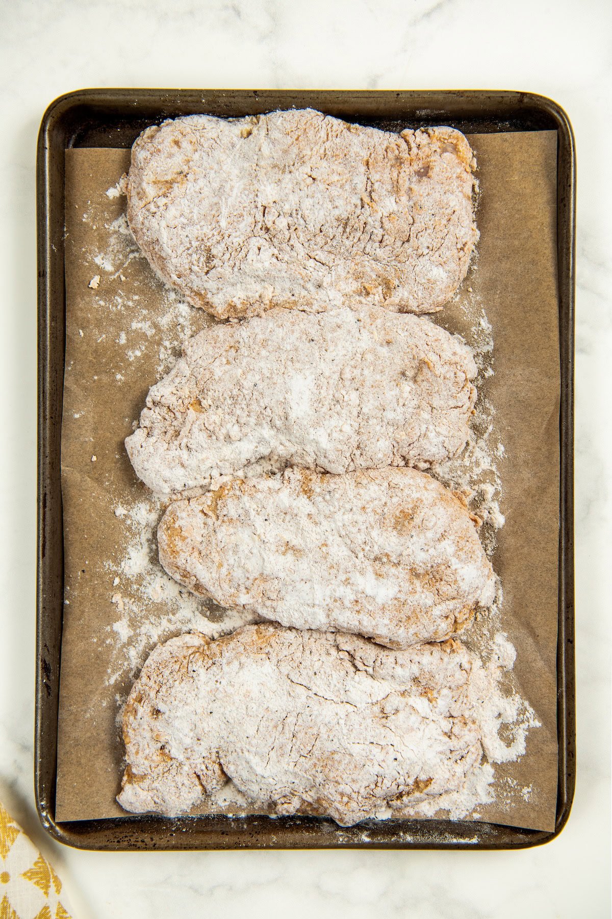 This Oven Fried Chicken recipe is always crispy, golden, perfectly juicy, and packed with flavor — without standing over a pot of hot oil. Four raw, breaded chicken cutlets covered in flour are arranged in a row on a parchment-lined baking sheet, ready to be cooked.