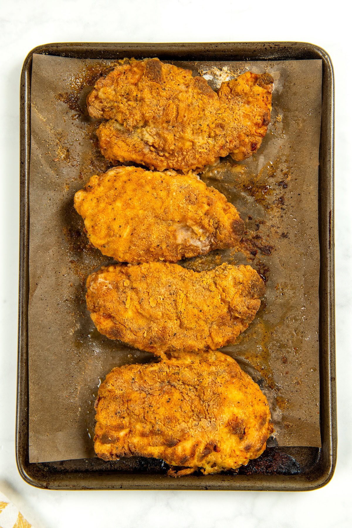This Oven Fried Chicken recipe is always crispy, golden, perfectly juicy, and packed with flavor — without standing over a pot of hot oil. Four golden-brown, breaded oven fried chicken breasts arranged in a row on a parchment-lined baking sheet.