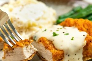 A close-up of oven fried chicken topped with creamy white gravy on a plate with mashed potatoes and green beans. A fork is holding a bite of the chicken.