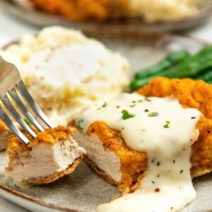 A close-up of oven fried chicken topped with creamy white gravy on a plate with mashed potatoes and green beans. A fork is holding a bite of the chicken.