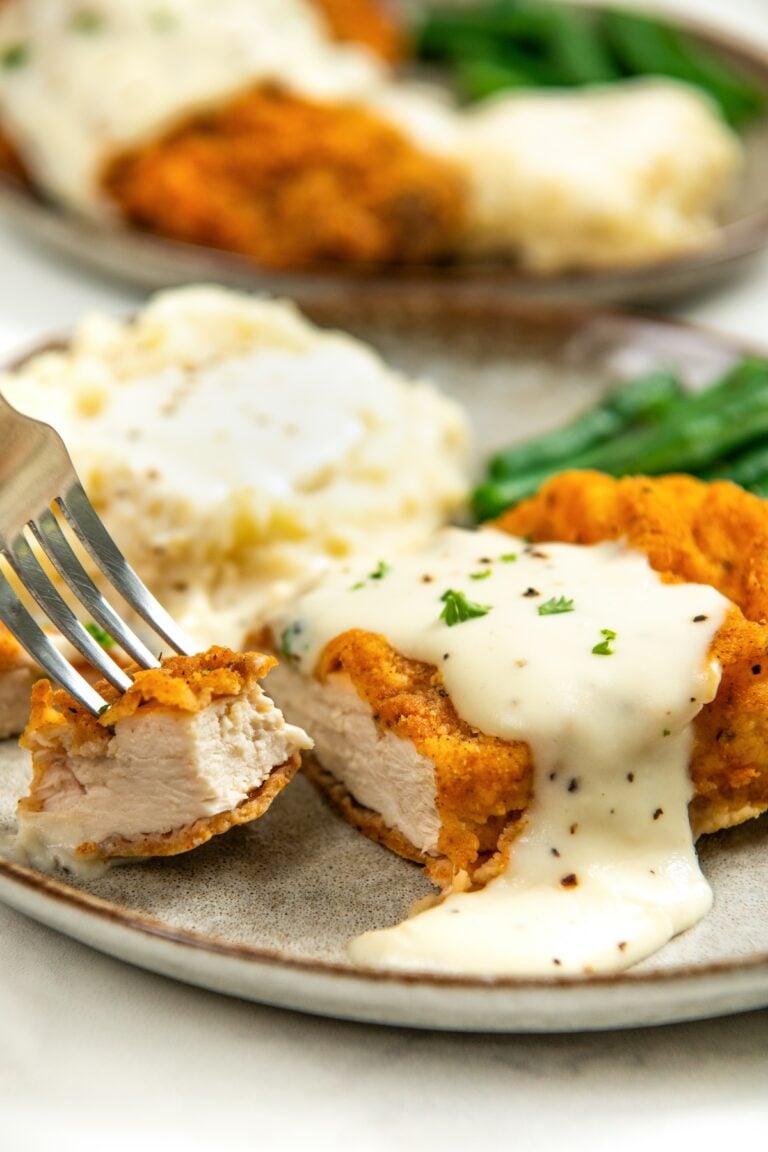 A close-up of oven fried chicken topped with creamy white gravy on a plate with mashed potatoes and green beans. A fork is holding a bite of the chicken.