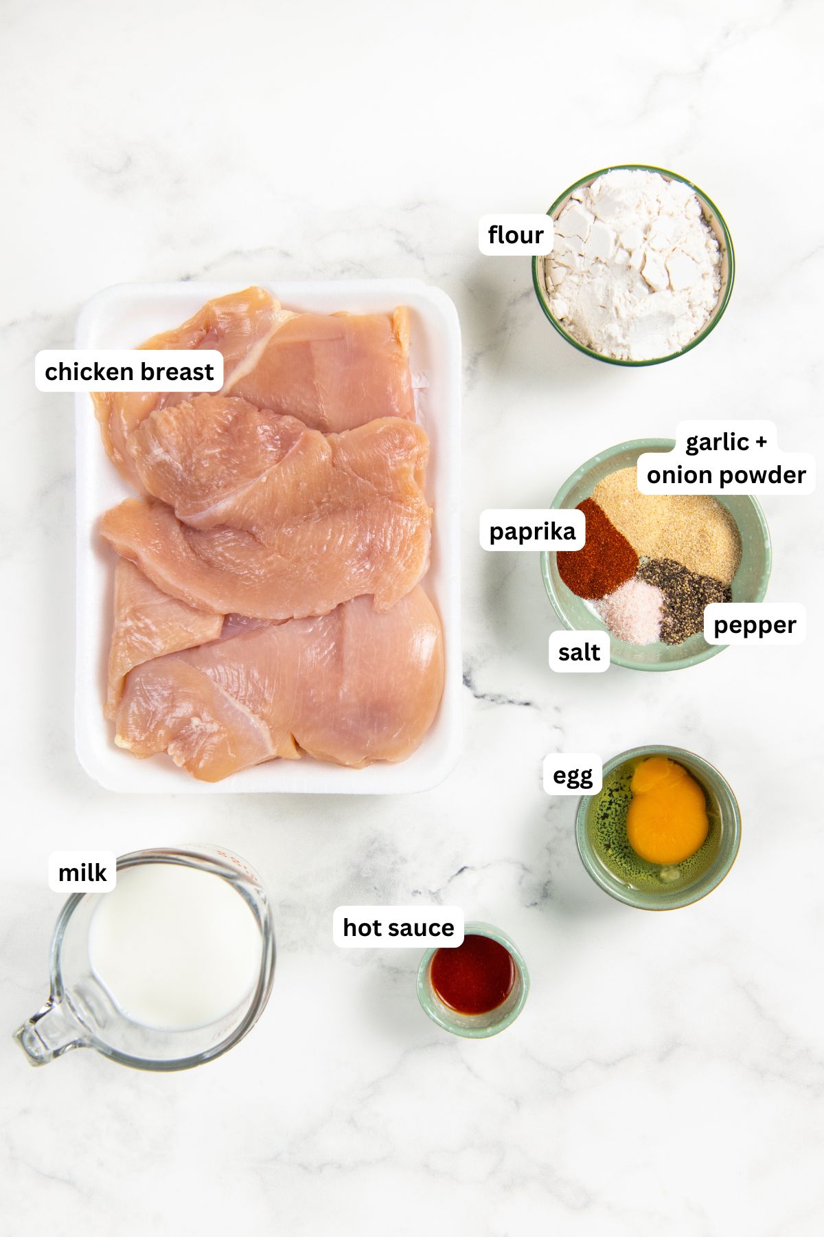This Oven Fried Chicken recipe is always crispy, golden, perfectly juicy, and packed with flavor — without standing over a pot of hot oil. Labeled ingredients for oven fried chicken recipe, from top to bottom: raw chicken breasts in a tray, with small bowls of flour, salt, black pepper, paprika, garlic and onion powder, a cup of milk, one egg, and hot sauce arranged on a white marble surface.