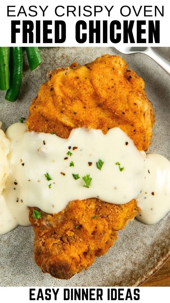 A breaded oven baked fried chicken breast topped with white gravy and chopped parsley, served with mashed potatoes and green beans on a gray plate.