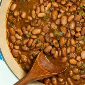 Learn how to cook pinto beans to tender and creamy perfection in a smoky, flavorful broth with bacon, jalapeño, onion, and more. A close-up of cooked pinto beans in a thick, brown sauce, mixed with jalapeños, in a blue pot. A wooden spoon rests in the beans, ready to serve.