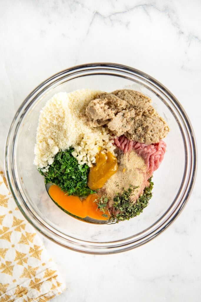 This easy Turkey Meatball recipe uses a few simple tricks and the perfect ingredient ratios to make moist, delicious meatballs every time! A glass bowl containing raw ground turkey, chopped herbs, minced garlic, breadcrumbs, grated cheese, chicken bouillon, and seasonings on a white marble surface.