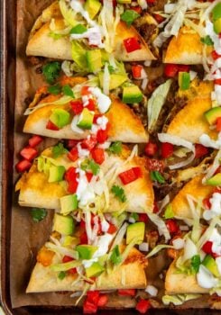 A baking sheet with baked tacos filled with ground beef, topped with shredded lettuce, diced avocado, tomato, green onions, cilantro, and drizzled with sour cream.
