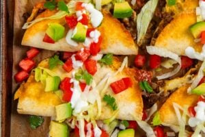 A baking sheet with baked tacos filled with ground beef, topped with shredded lettuce, diced avocado, tomato, green onions, cilantro, and drizzled with sour cream.