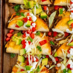A baking sheet with baked tacos filled with ground beef, topped with shredded lettuce, diced avocado, tomato, green onions, cilantro, and drizzled with sour cream.