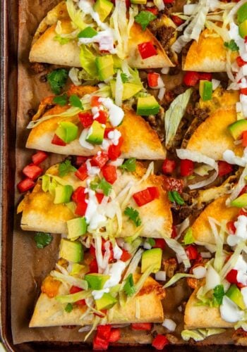 A baking sheet with baked tacos filled with ground beef, topped with shredded lettuce, diced avocado, tomato, green onions, cilantro, and drizzled with sour cream.