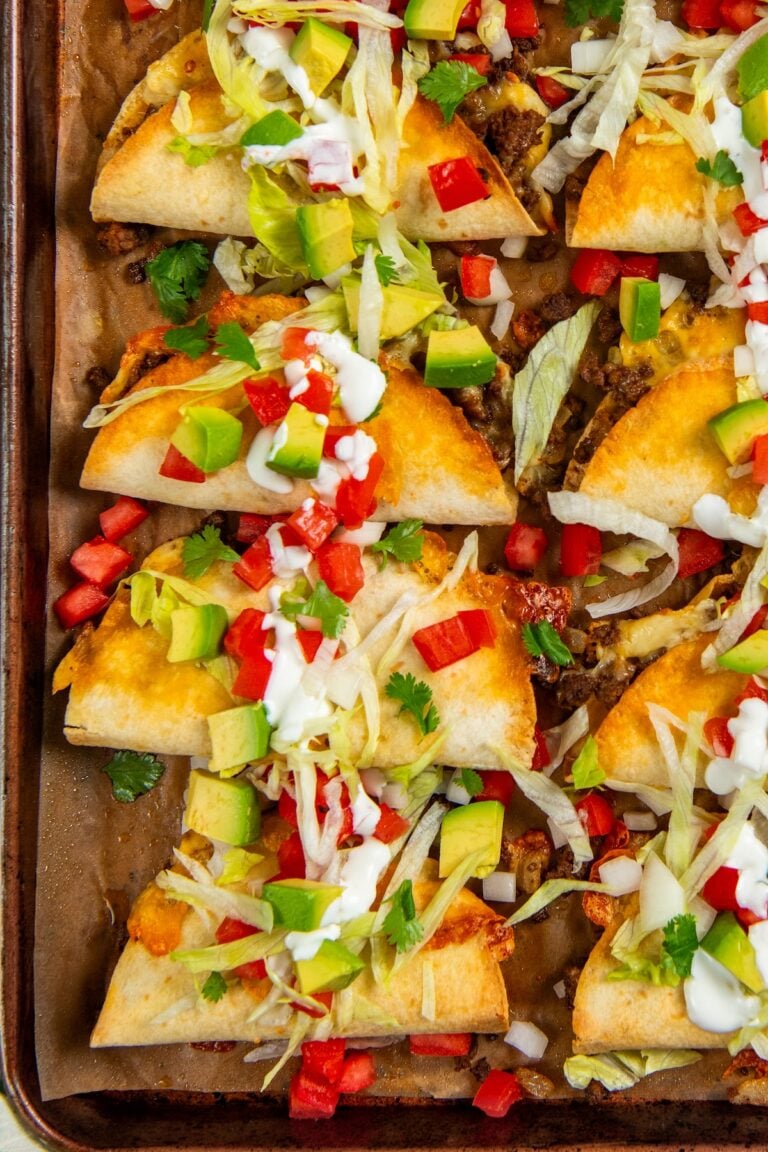 A baking sheet with baked tacos filled with ground beef, topped with shredded lettuce, diced avocado, tomato, green onions, cilantro, and drizzled with sour cream.