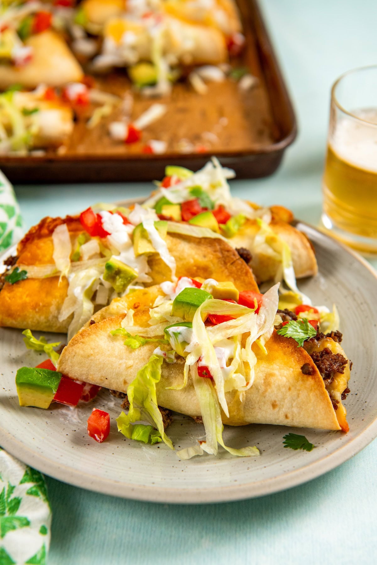 Sheet Pan Tacos are the easiest way to get crispy, cheesy, restaurant-worthy baked tacos on the table ready to serve all at the same time! Three baked sheet pan tacos topped with shredded lettuce, diced avocado, and chopped red peppers on a plate, with a baking sheet of more tacos and a glass of beer in the background.