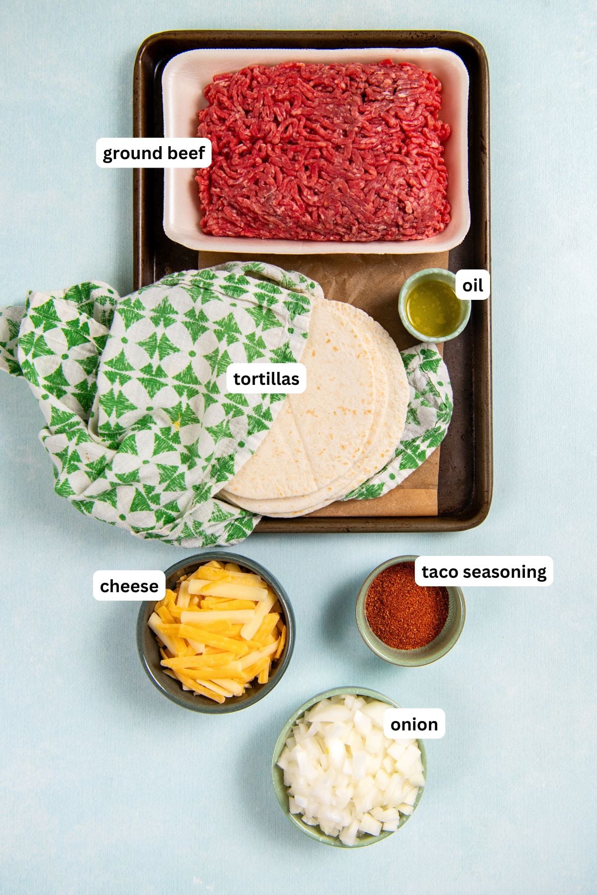 Sheet Pan Tacos are the easiest way to get crispy, cheesy, restaurant-worthy baked tacos on the table ready to serve all at the same time! Labeled ingredients for sheet pan tacos: ground beef, a stack of tortillas, a small dish of oil, shredded cheese, taco seasoning, and chopped onion.