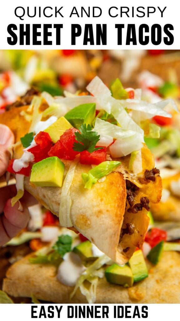 A close-up of a hand holding a crispy sheet pan taco topped with diced avocado, lettuce, tomato, cilantro, and sour cream, with more tacos in the background.