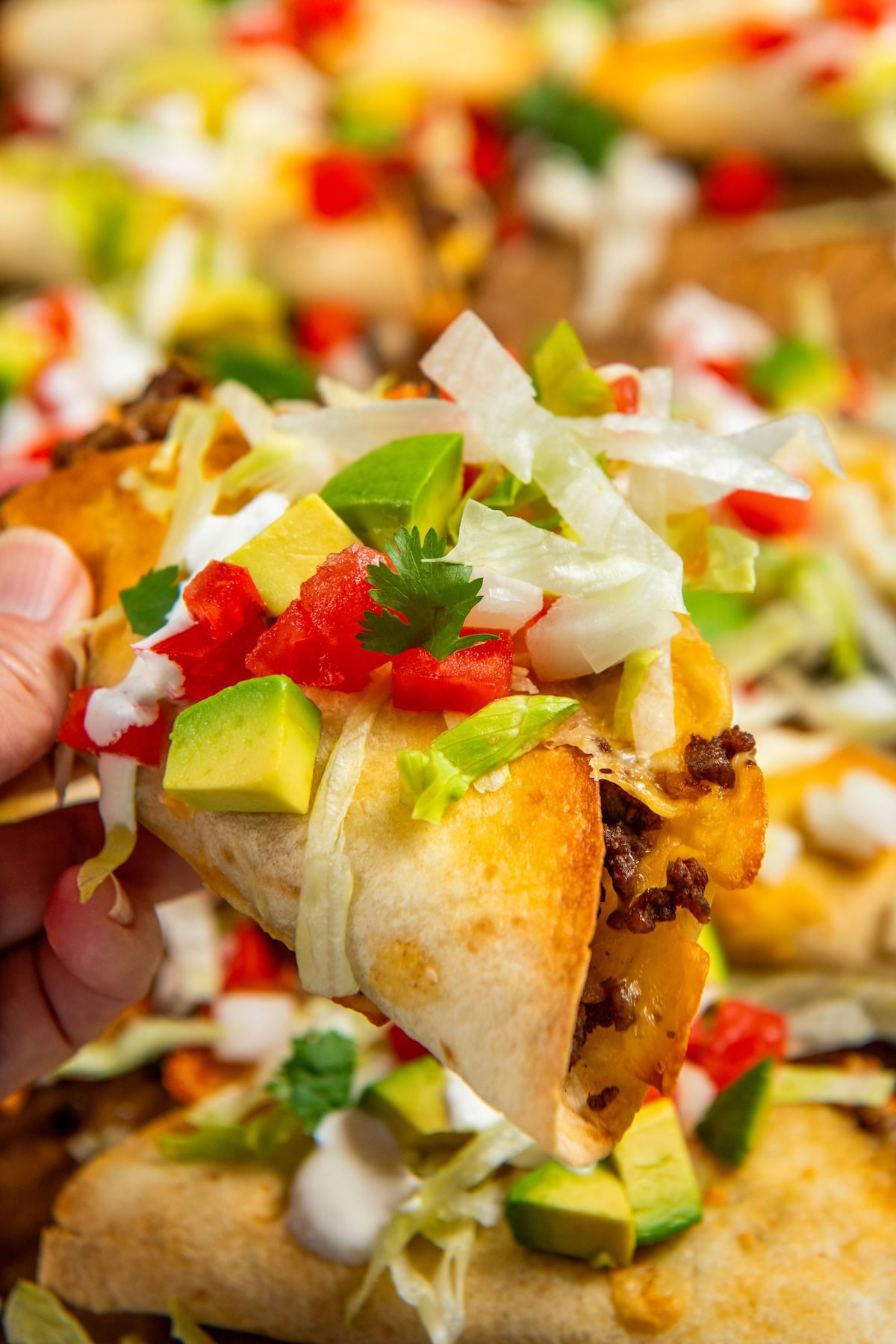 Sheet Pan Tacos are the easiest way to get crispy, cheesy, restaurant-worthy baked tacos on the table ready to serve all at the same time! A hand holding a crispy baked taco filled with ground meat, melted cheese, diced avocado, chopped tomatoes, shredded lettuce, cilantro, and drizzled with a white sauce. Other tacos on a sheet pan are blurred in the background.